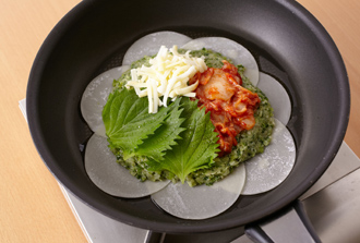 端を少し空けてひき肉だねを平らにのせ、青じそ、白菜キムチ、ピザ用チーズをのせる。下に敷いた餃子の皮の端に薄く水を塗り、さらに皮7枚をドーナツ状に丸くのせて端を閉じ、中央に1枚をのせる。